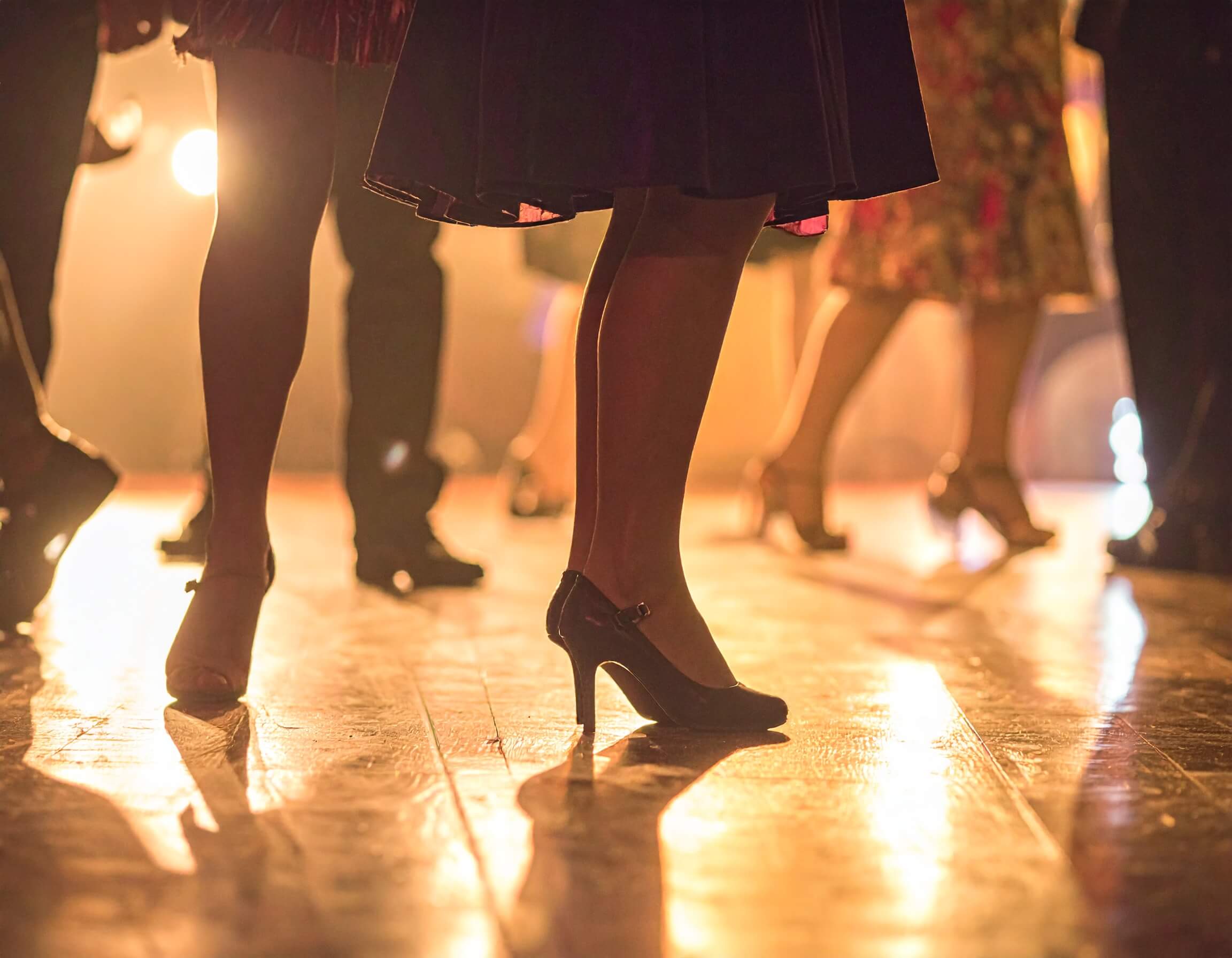 Eine belebte Tanzfläche in einem Saal. Der Fokus liegt auf den Beinen und Füßen der tanzenden Personen.