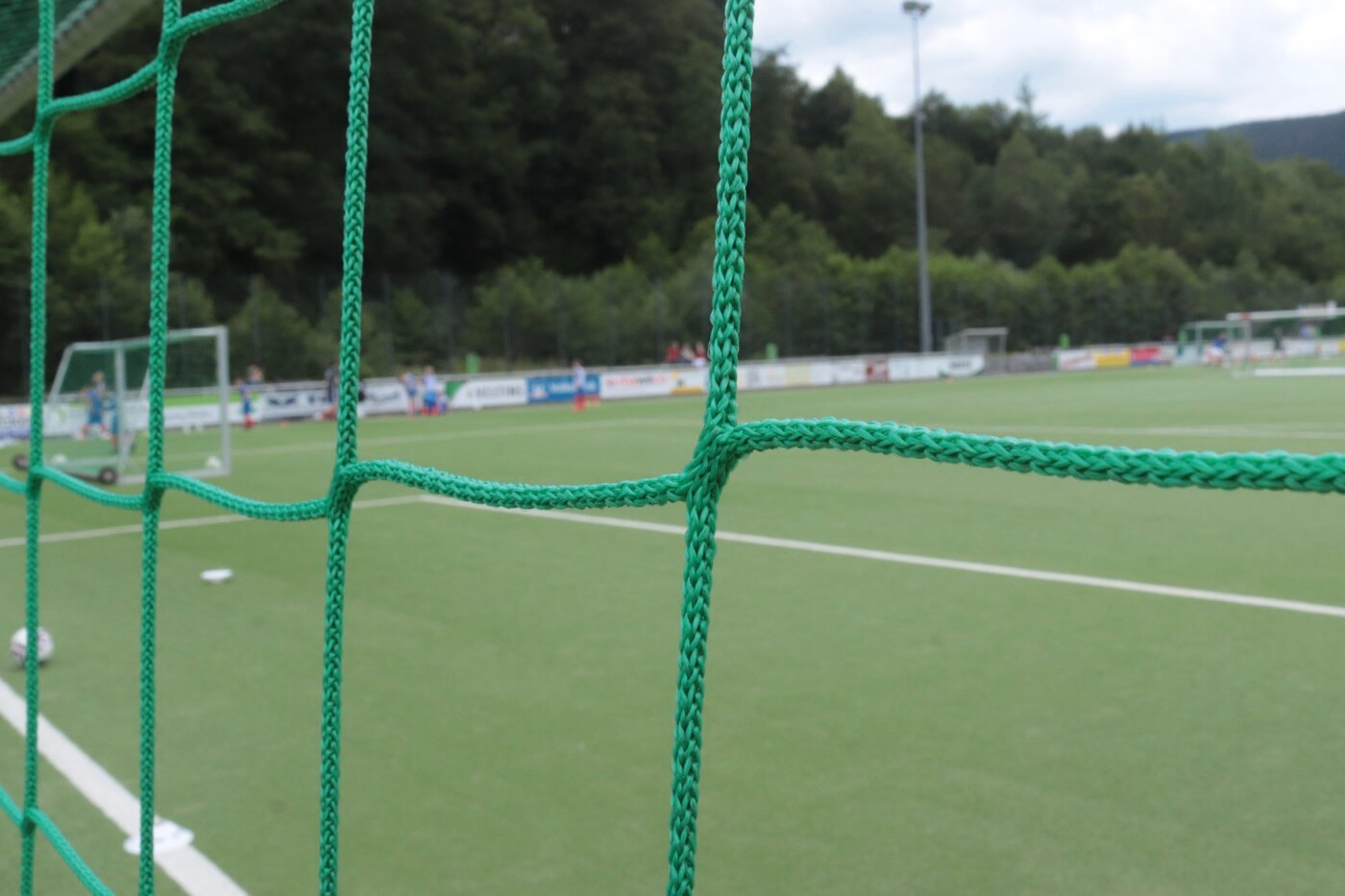 Nahaufnahme eines Tornetzes, durch das Tornetz sieht man unscharf einen Fussballplatz.