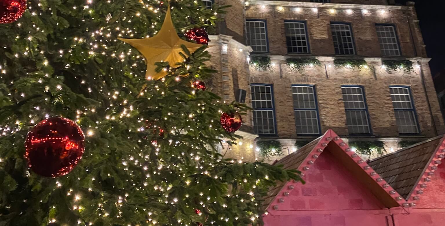 Links im Bild ist ein festlich geschmückter Weihnachtsbaum, im Hintergrund ein älteres Haus