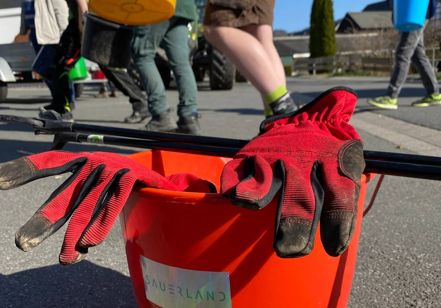 Nahaufnahme eines Eimer mit einem newsauerland Aufkleber. Im Hintergrund sieht man viele Beine die los gehen um die Landschaft sauberer zu machen.