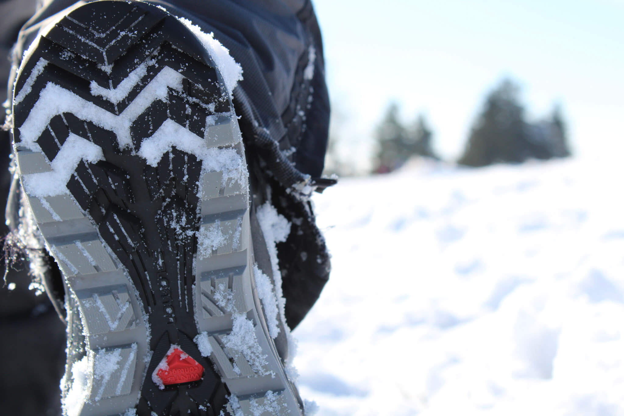 Die Sohle eines Wanderschuhs zeigt in die Kamera, ein Wanderer wandert durch eine Schneebedeckte Landschaft